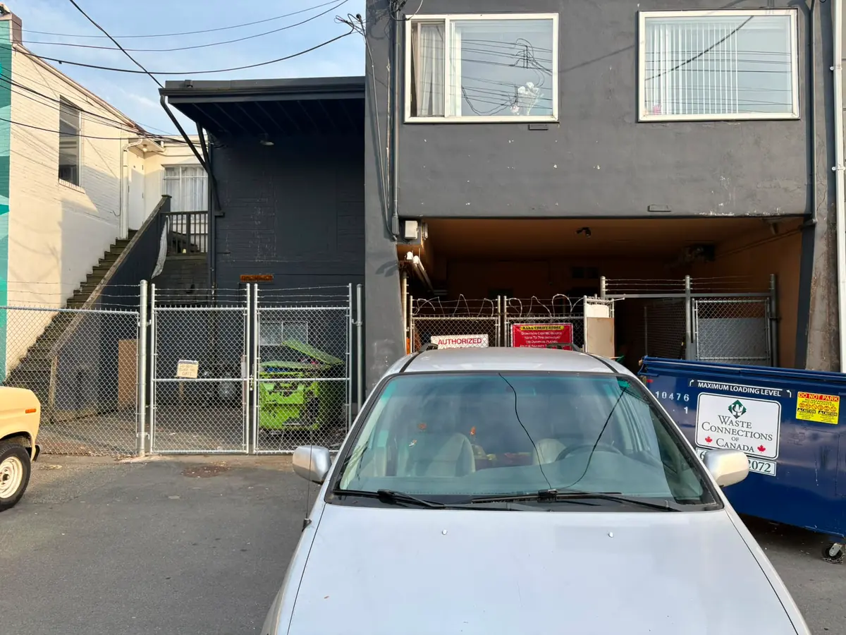 The lane behind Raba Thrift Store, showing the chain-link fence with the red Raba Thrift Store donation centre sign, a blue waste bin, and the covered bay used for donation drop-off.