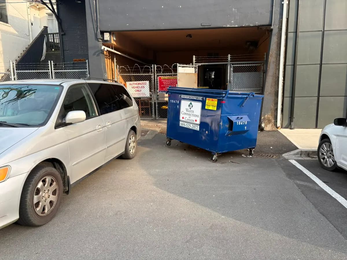 Closer view of the donation drop-off area behind Raba Thrift Store: open paved space beside a blue waste bin, with the covered donation bay straight ahead.
